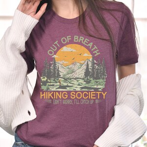 May include: A maroon t-shirt with a vintage-style graphic of a mountain range with a hiker and the text "Out of Breath Hiking Society Don't Worry, I'll Catch Up."