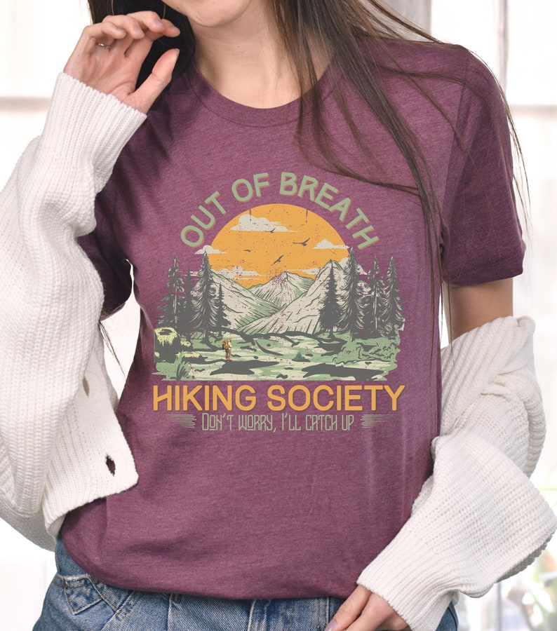 May include: A maroon t-shirt with a vintage-style graphic of a mountain range with a hiker and the text "Out of Breath Hiking Society Don't Worry, I'll Catch Up."