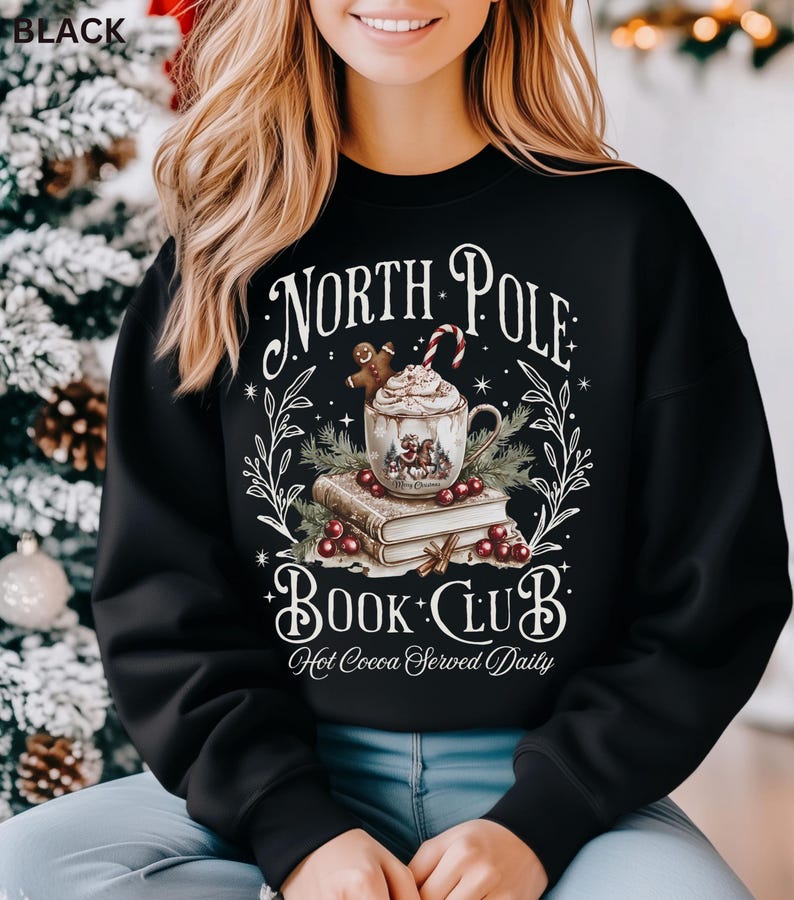 Puede incluir: Sudadera negra con un dise&ntilde;o festivo. El gr&aacute;fico presenta una taza de chocolate caliente, un hombre de jengibre y un bast&oacute;n de caramelo, con el texto "North Pole Book Club: Hot Cocoa Served Daily". El dise&ntilde;o est&aacute; rodeado de hojas y bayas.