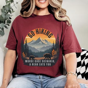 May include: A maroon t-shirt with a graphic of a mountain range and the text "Go Hiking" in a circle. Below the circle is the text "Worse case scenario, a bear eats you".