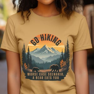 May include: A mustard yellow t-shirt with a graphic of a mountain range and two bears. The graphic is in a circle with the text "Go Hiking" above the mountain range and "Worse case scenario, a bear eats you" below the mountain range.