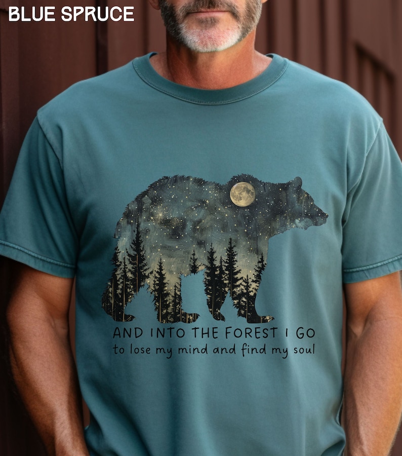 Pu&ograve; includere: Una t-shirt blu turchese con una grafica di un orso in silhouette con un cielo notturno stellato e una foresta al suo interno. Il testo "AND INTO THE FOREST I GO to lose my mind and find my soul" &egrave; stampato sotto la grafica.