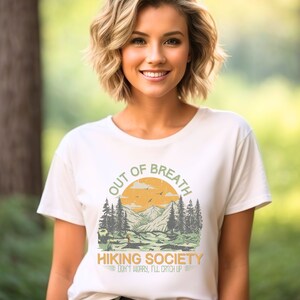 May include: White t-shirt with a graphic of a mountain landscape and the text "Out of Breath Hiking Society Don't worry, I'll catch up."