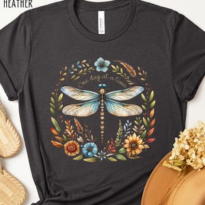 May include: Dark grey heather t-shirt with a floral design featuring a dragonfly and the text "One day at a time".