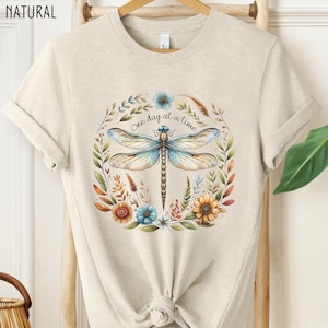 May include: A light beige t-shirt with a floral design featuring a dragonfly and the text "One day at a time".