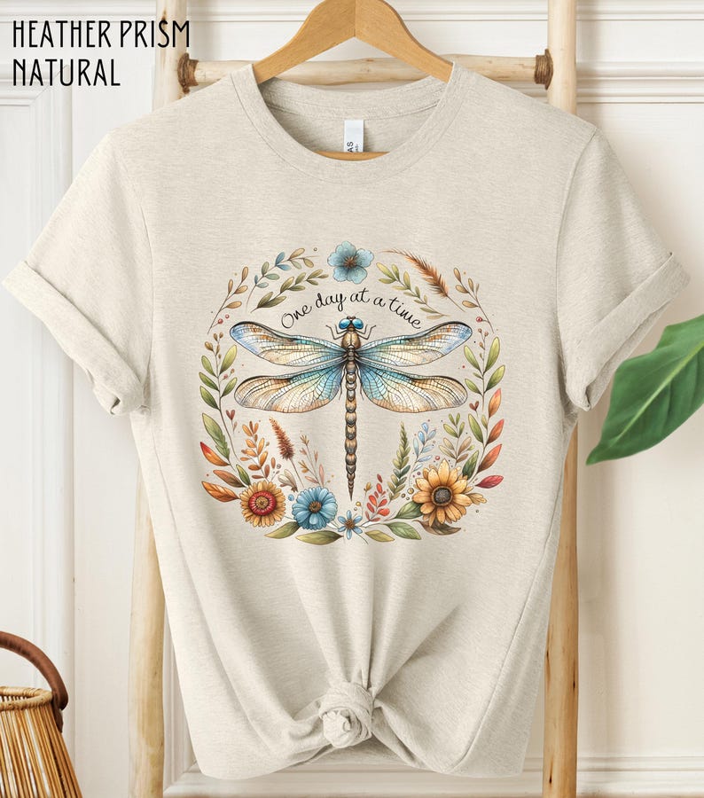 May include: A light beige t-shirt with a floral design featuring a dragonfly and the text "One day at a time".