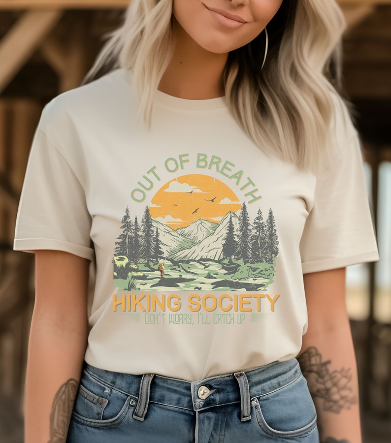 May include: A white t-shirt with a vintage-style graphic of a mountain range with a sunset and the text "Out of Breath Hiking Society Don't Worry, I'll Catch Up".