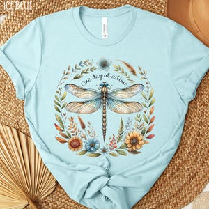 May include: A light blue t-shirt with a dragonfly and floral design. The text "One day at a time" is printed on the shirt.