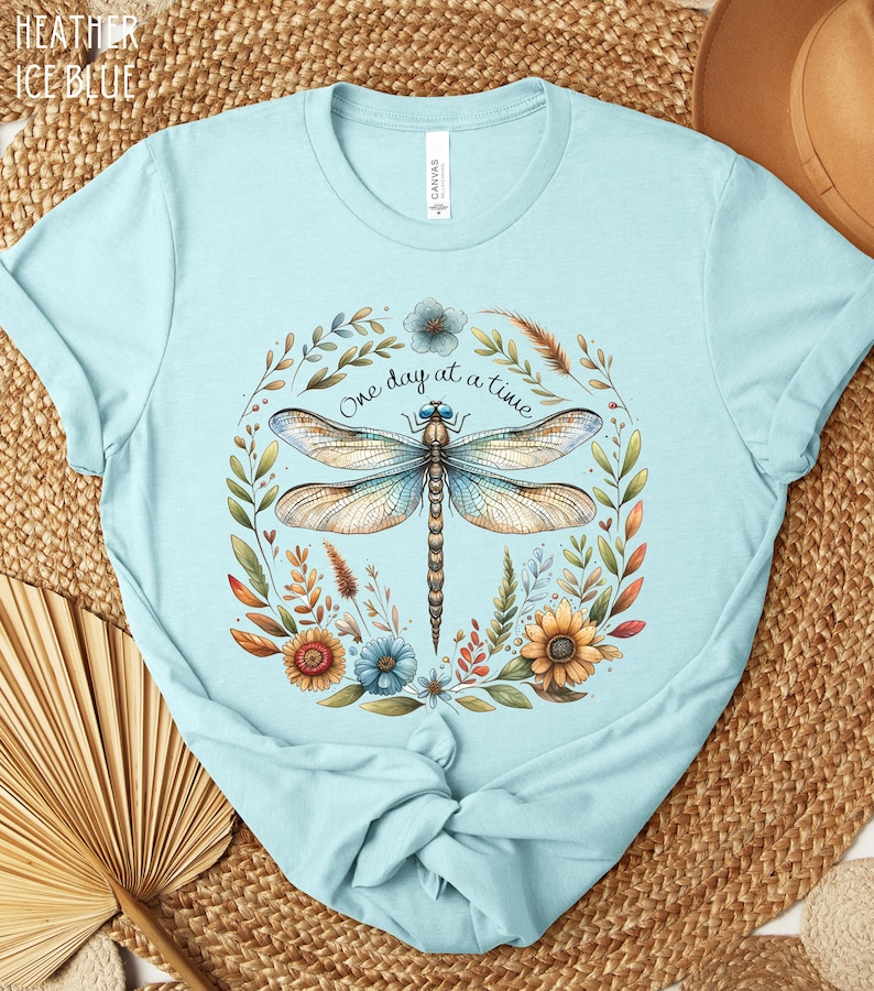 May include: A light blue t-shirt with a dragonfly and floral design. The text "One day at a time" is printed on the shirt.