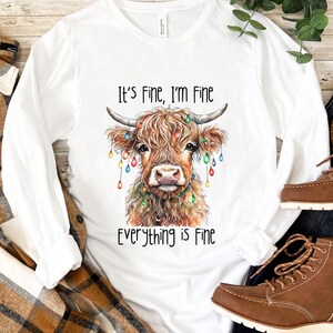 Puede incluir: Camiseta blanca de manga larga con una ilustración de acuarela de una vaca de las Tierras Altas adornada con luces navideñas de colores. El texto "It's Fine, I'm Fine, Everything is Fine" está impreso debajo.