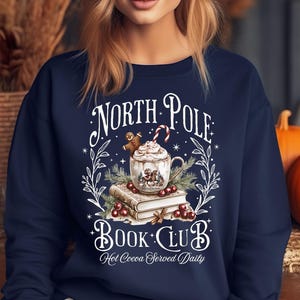 Puede incluir: Sudadera azul marino con un dise&ntilde;o festivo. El gr&aacute;fico presenta una taza de chocolate caliente con crema batida, un hombre de jengibre y un bast&oacute;n de caramelo. El texto dice "North Pole Book Club" y "Hot Cocoa Served Daily".