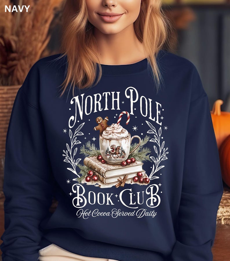 Puede incluir: Sudadera azul marino con un dise&ntilde;o festivo. El gr&aacute;fico presenta una taza de chocolate caliente con crema batida, un hombre de jengibre y un bast&oacute;n de caramelo. El texto dice "North Pole Book Club" y "Hot Cocoa Served Daily".