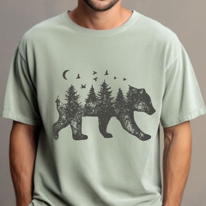 May include: A light green t-shirt with a black silhouette of a bear. The bear is made up of trees and birds. The design is centered on the shirt.