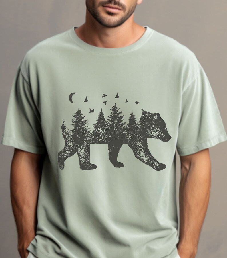 May include: A light green t-shirt with a black silhouette of a bear. The bear is made up of trees and birds. The design is centered on the shirt.