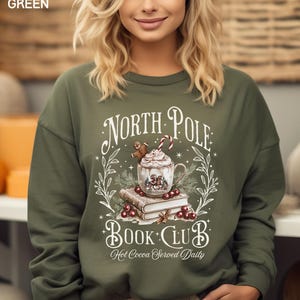 Puede incluir: Sudadera verde militar con la inscripci&oacute;n "North Pole Book Club" y "Hot Cocoa Served Daily". El dise&ntilde;o incluye una taza de chocolate caliente, un hombre de jengibre, un bast&oacute;n de caramelo y libros. La sudadera es de cuello redondo.