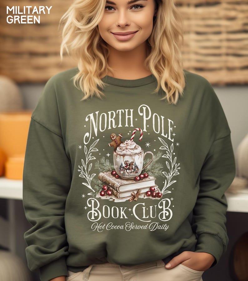 Puede incluir: Sudadera verde militar con la inscripci&oacute;n "North Pole Book Club" y "Hot Cocoa Served Daily". El dise&ntilde;o incluye una taza de chocolate caliente, un hombre de jengibre, un bast&oacute;n de caramelo y libros. La sudadera es de cuello redondo.