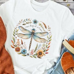 May include: White t-shirt with a floral wreath design featuring a dragonfly and the text "One day at a time".