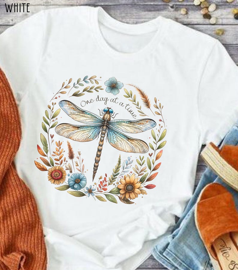 May include: White t-shirt with a floral wreath design featuring a dragonfly and the text "One day at a time".