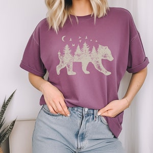 May include: A purple short-sleeved t-shirt with a graphic of a bear silhouette made up of trees and a crescent moon. The bear is facing right and there are birds flying above the trees.