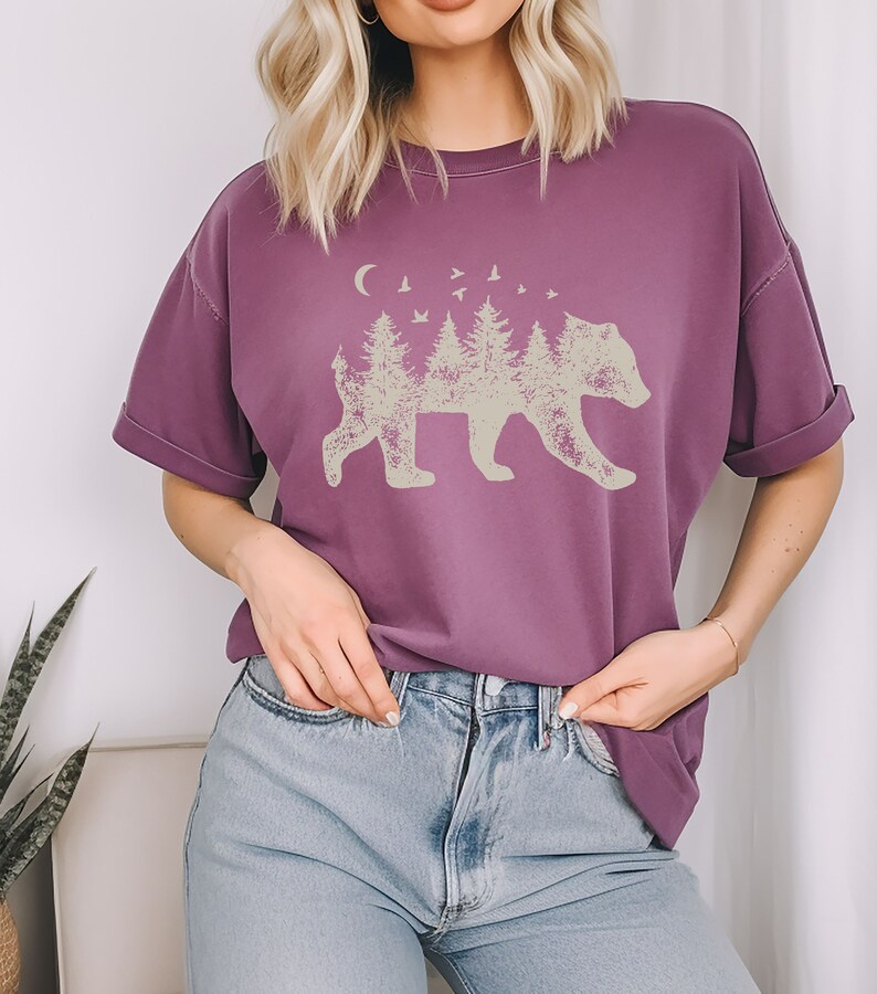 May include: A purple short-sleeved t-shirt with a graphic of a bear silhouette made up of trees and a crescent moon. The bear is facing right and there are birds flying above the trees.