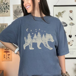 May include: A blue t-shirt with a graphic of a bear silhouette made of trees and a crescent moon with birds flying above.