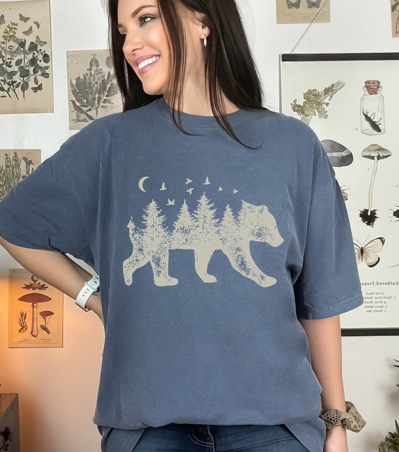 May include: A blue t-shirt with a graphic of a bear silhouette made of trees and a crescent moon with birds flying above.
