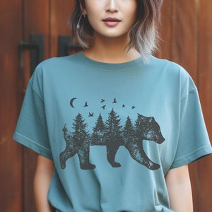 May include: A teal blue t-shirt with a black bear silhouette design. The bear is made up of trees and birds, with a crescent moon above.