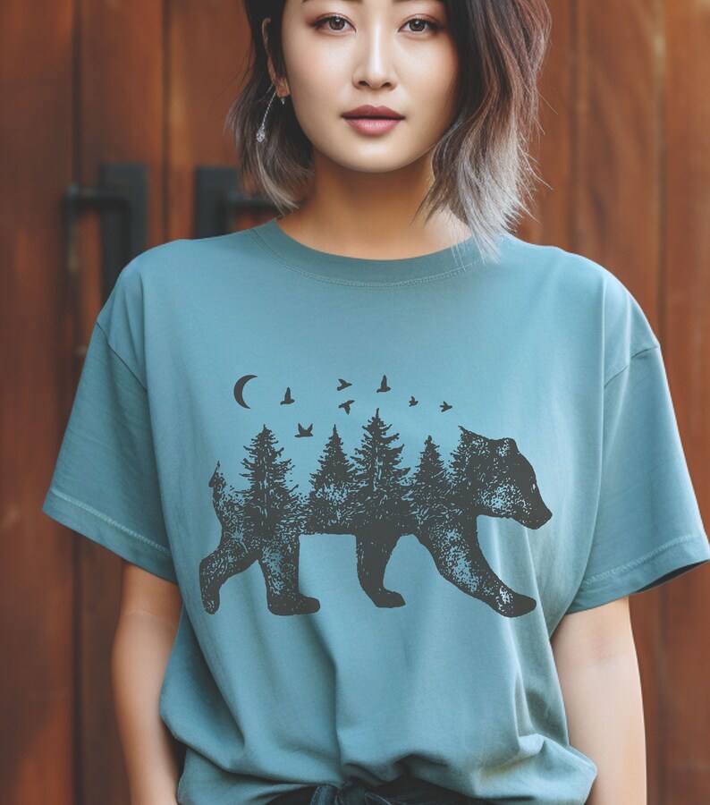 May include: A teal blue t-shirt with a black bear silhouette design. The bear is made up of trees and birds, with a crescent moon above.