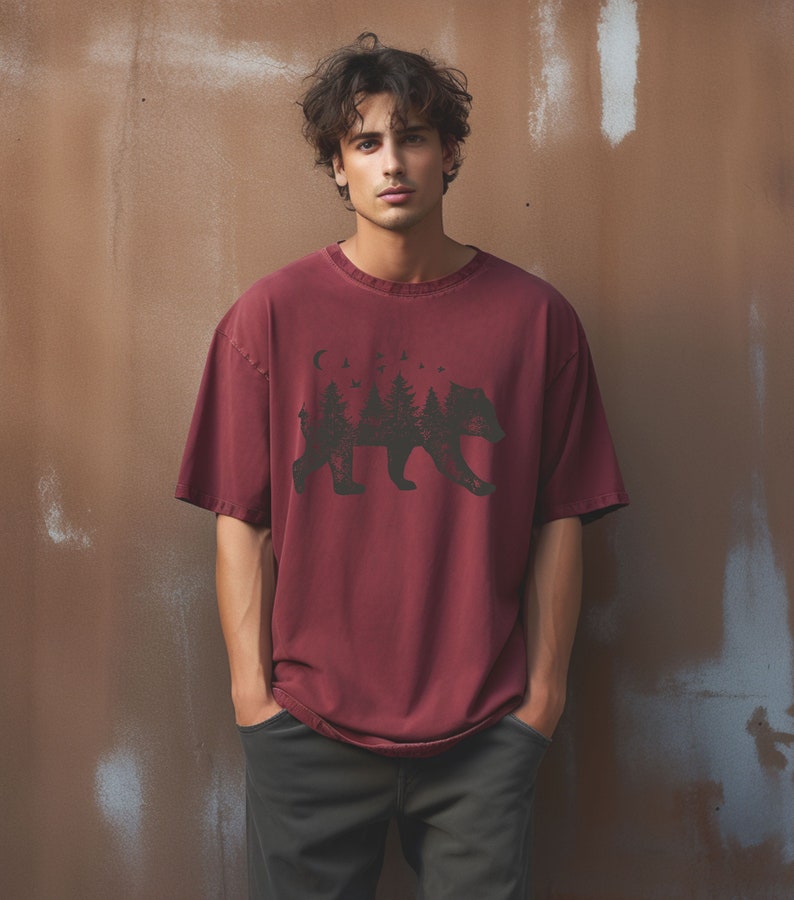 May include: A maroon t-shirt with a black graphic of a bear silhouette with trees and a crescent moon inside the bear's body.