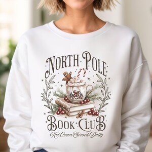 Puede incluir: Sudadera blanca con un dise&ntilde;o de "North Pole Book Club" que muestra una taza de chocolate caliente, un hombre de jengibre, un bast&oacute;n de caramelo y libros. Tambi&eacute;n incluye la frase "Hot Cocoa Served Daily".