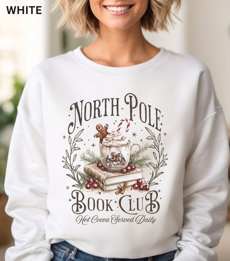 Puede incluir: Sudadera blanca con un dise&ntilde;o de "North Pole Book Club" que muestra una taza de chocolate caliente, un hombre de jengibre, un bast&oacute;n de caramelo y libros. Tambi&eacute;n incluye la frase "Hot Cocoa Served Daily".