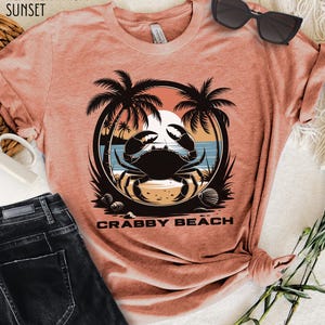 May include: A heather prism sunset colored t-shirt with a graphic of a crab on a beach with palm trees and the text "Crabby Beach".