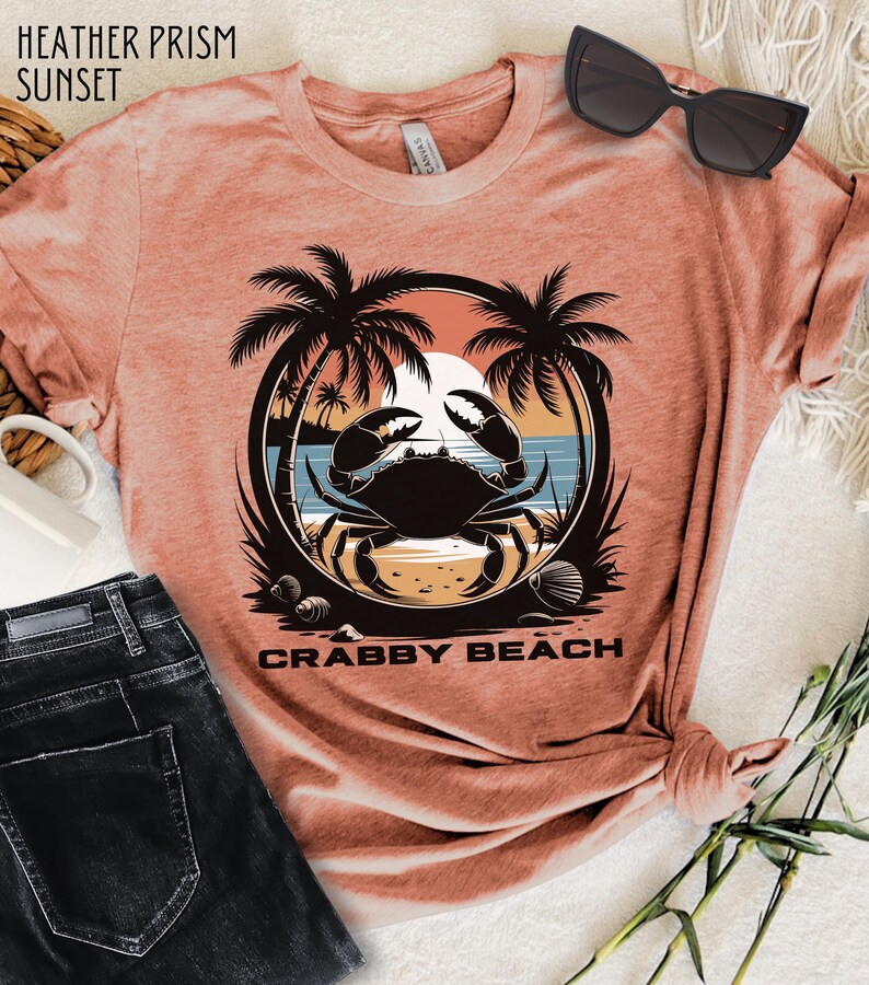 May include: A heather prism sunset colored t-shirt with a graphic of a crab on a beach with palm trees and the text "Crabby Beach".