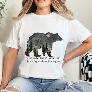Pu&ograve; includere: Maglietta bianca con una grafica di un orso in silhouette con una luna e alberi al suo interno. Il testo "AND INTO THE FOREST I GO to lose my mind and find my soul" &egrave; stampato sotto l'orso.