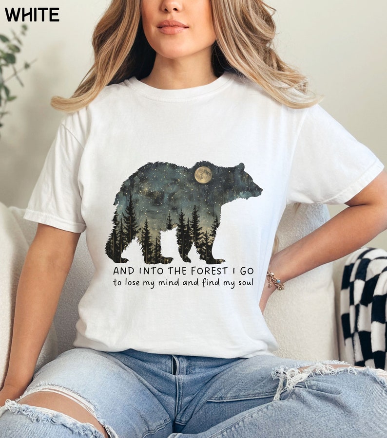 Pu&ograve; includere: Maglietta bianca con una grafica di un orso in silhouette con una luna e alberi al suo interno. Il testo "AND INTO THE FOREST I GO to lose my mind and find my soul" &egrave; stampato sotto l'orso.
