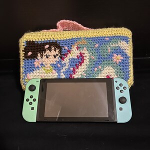 May include: A crocheted case with a floral design and a cartoon character. The case is light yellow, blue, green, pink, and white. A teal Nintendo Switch console is in front of the case.