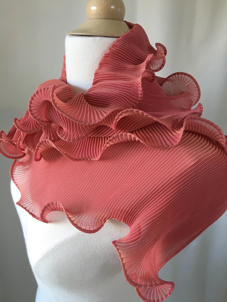 May include: A coral pink ruffled scarf made of pleated fabric. The scarf is draped around a white mannequin's neck.