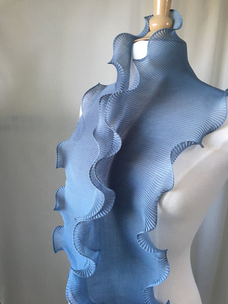 May include: A light blue pleated fabric scarf with a ruffled edge. The scarf is draped over a white dress form.