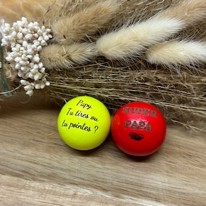 Peut inclure: Deux boules en bois avec du texte. La boule jaune dit "Papy, Tu tires ou tu pointes?" et la boule rouge dit "Super Papa".