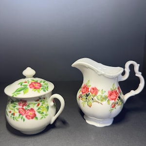 Può includere: Zuccheriera in ceramica bianca con coperchio e una lattiera, entrambe decorate con un motivo floreale di rose rosse, verdi e bianche. La zuccheriera ha un manico e la lattiera ha un manico e un beccuccio curvi.