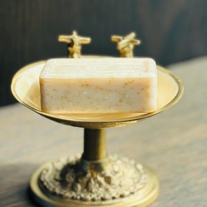 May include: A rectangular bar of off-white soap with visible brown flecks resting on a gold-toned metal pedestal dish. The dish features a circular base with ornate detailing and two small cross-shaped handles extending from the back rim.