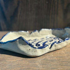 May include: A handmade ceramic dish with a unique, organic shape. The dish features a light beige interior with blue decorative patterns and a dark blue rim. The textured surface adds to its rustic charm. Perfect for serving snacks or as a decorative piece.