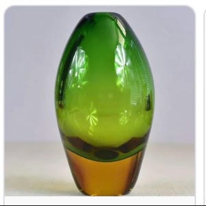 May include: A smooth, egg-shaped glass vase featuring a vibrant emerald green upper section that transitions into a solid amber-coloured base. The glass is translucent, reflecting light with a polished finish and a rounded, tapered silhouette.