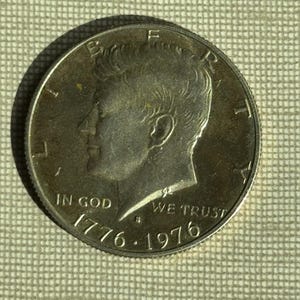 Op de afbeelding: Een zilveren munt met een profiel van een man, met de inscripties "LIBERTY", "IN GOD WE TRUST" en de data "1776-1976". De munt heeft een gedetailleerde rand, waarschijnlijk een herdenkings-halve dollar.