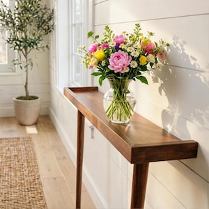 Op de afbeelding: Een houten console met een vaas vol kleurrijke bloemen. De vaas bevat roze pioenrozen, gele en witte madeliefjes en andere bloemen. De tafel heeft een lange, smalle bovenkant en schuine poten. Op de achtergrond staat een olijfboom in een pot.