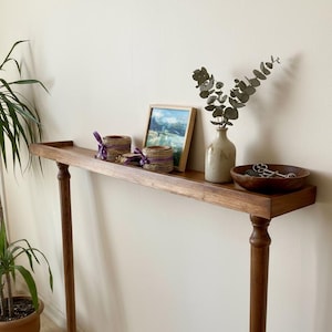 May include: A long, narrow, wooden console table with turned legs. The tabletop displays a framed landscape painting, two woven candle holders with purple ribbon, a small vase with greenery, and a wooden bowl with keys.