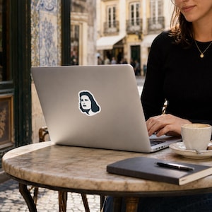 Op de afbeelding: Een laptop met een sticker van een vrouwengezicht in marineblauw en wit. De laptop staat op een ronde tafel met een marmeren blad. Een koffiekopje en een notitieboek liggen ook op de tafel. De achtergrond toont een straatbeeld met gebouwen en een blauw-witte tegelmuur.
