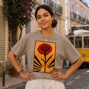 Op de afbeelding: Een vrouw draagt een grijs t-shirt met een afbeelding van een rode bloem op een oranje achtergrond. Het bloemontwerp is gecentreerd op het shirt. De vrouw staat buiten voor een gebouw en een gele tram.