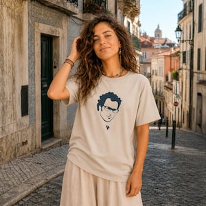 Op de afbeelding: Beige T-shirt met een marineblauwe illustratie van een gezicht met een bril en krullend haar. Het T-shirt wordt gedragen met een beige broek. De achtergrond toont een geplaveide straat en gebouwen.