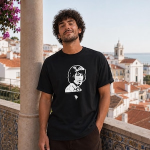 Op de afbeelding: Zwart t-shirt met een witte portretafbeelding. Het t-shirt heeft een ronde hals en korte mouwen. De persoon op de foto draagt het t-shirt. De achtergrond toont een stadsgezicht met gebouwen en een watermassa.